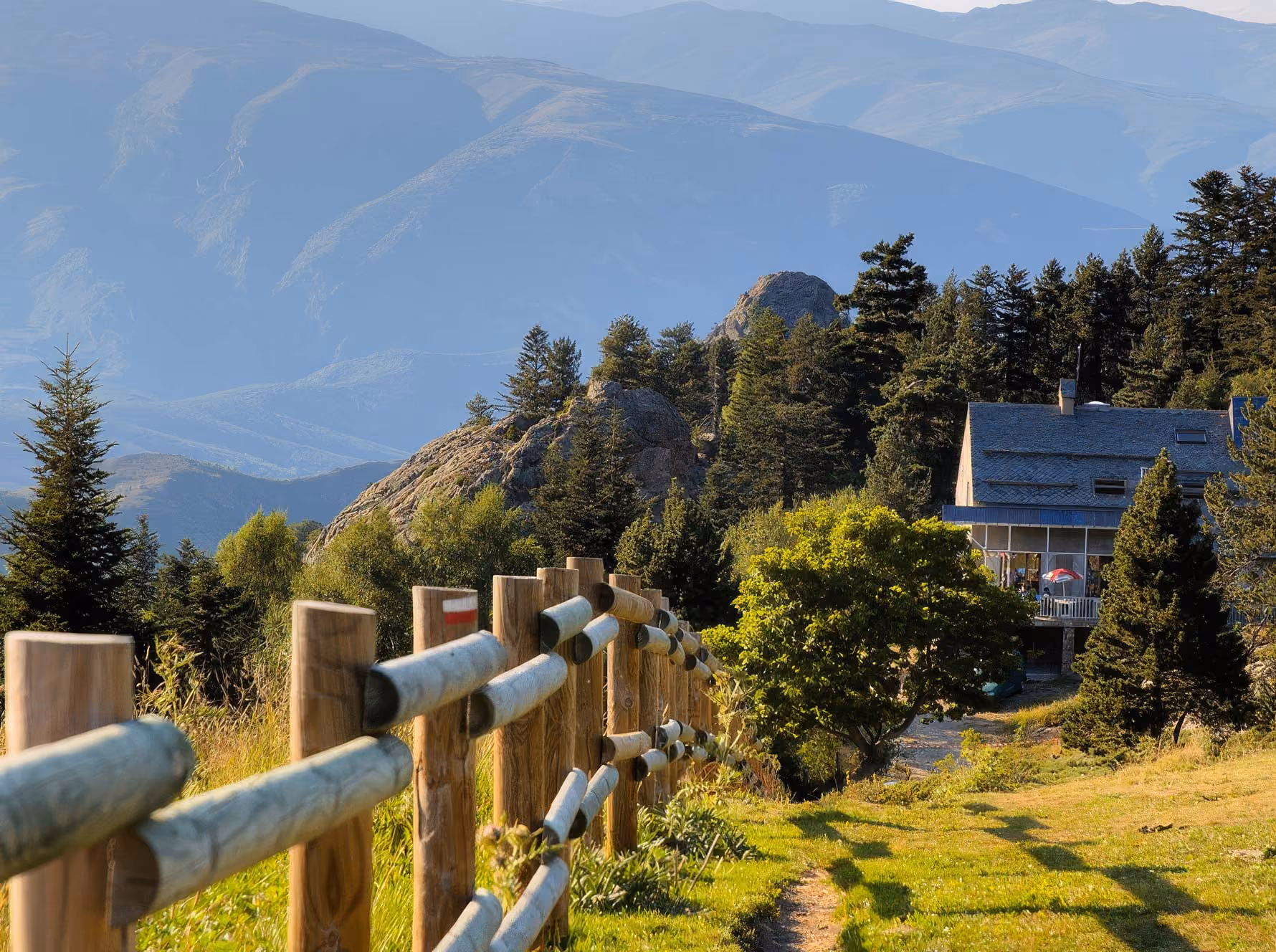 Wooden fence leading towards a house surrounded by trees on a grassy mountain slope, with layered blue mountains visible in the background.
