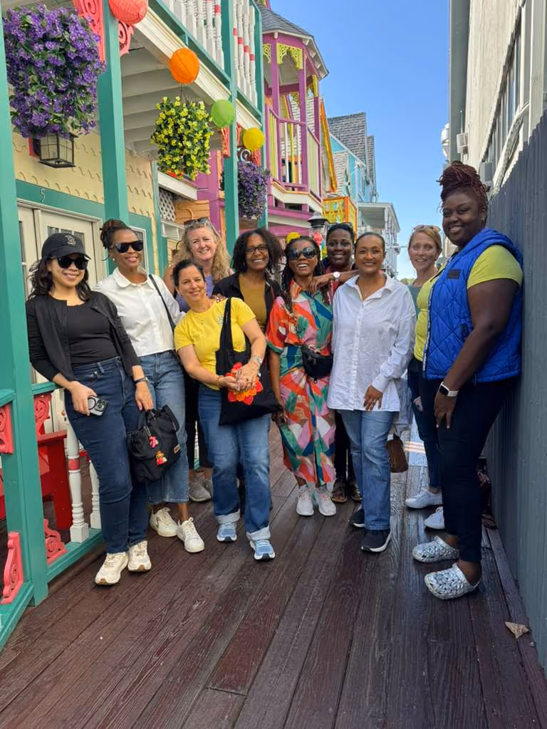 Group of nine women standing on a wooden walkway between colorful buildings decorated with hanging flowers and lanterns, smiling at the camera on a sunny day.