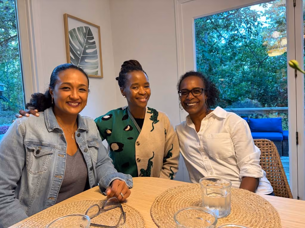 Three women sitting closely at a wooden table indoors, smiling warmly at the camera. The background shows a framed leaf print on the wall and large windows revealing green trees outside.
