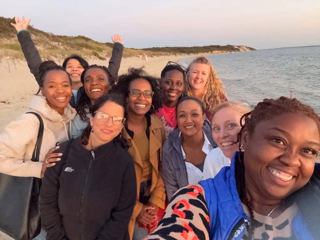 Group of ten diverse women smiling and posing for a selfie on a sandy beach with dunes and calm ocean in the background during sunset.