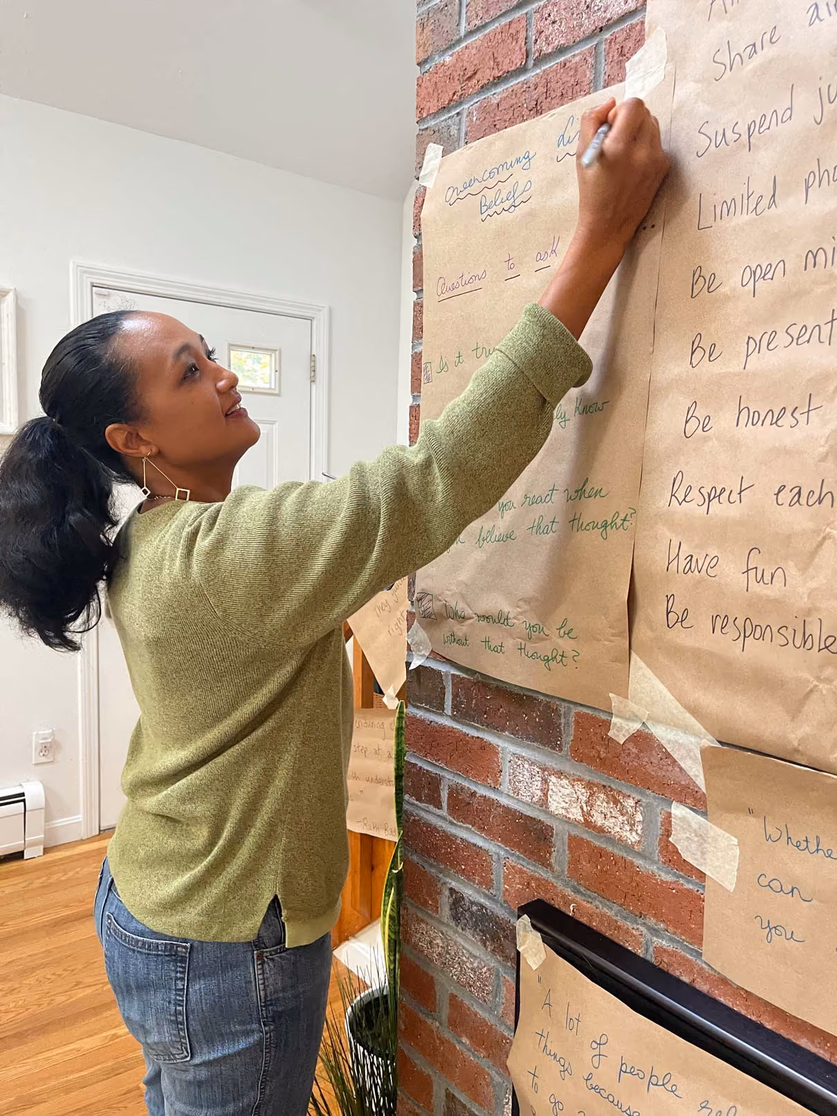 Clementine writes with a marker on taped large brown paper sheets affixed to a red brick wall in a room with hardwood floors and a white door.