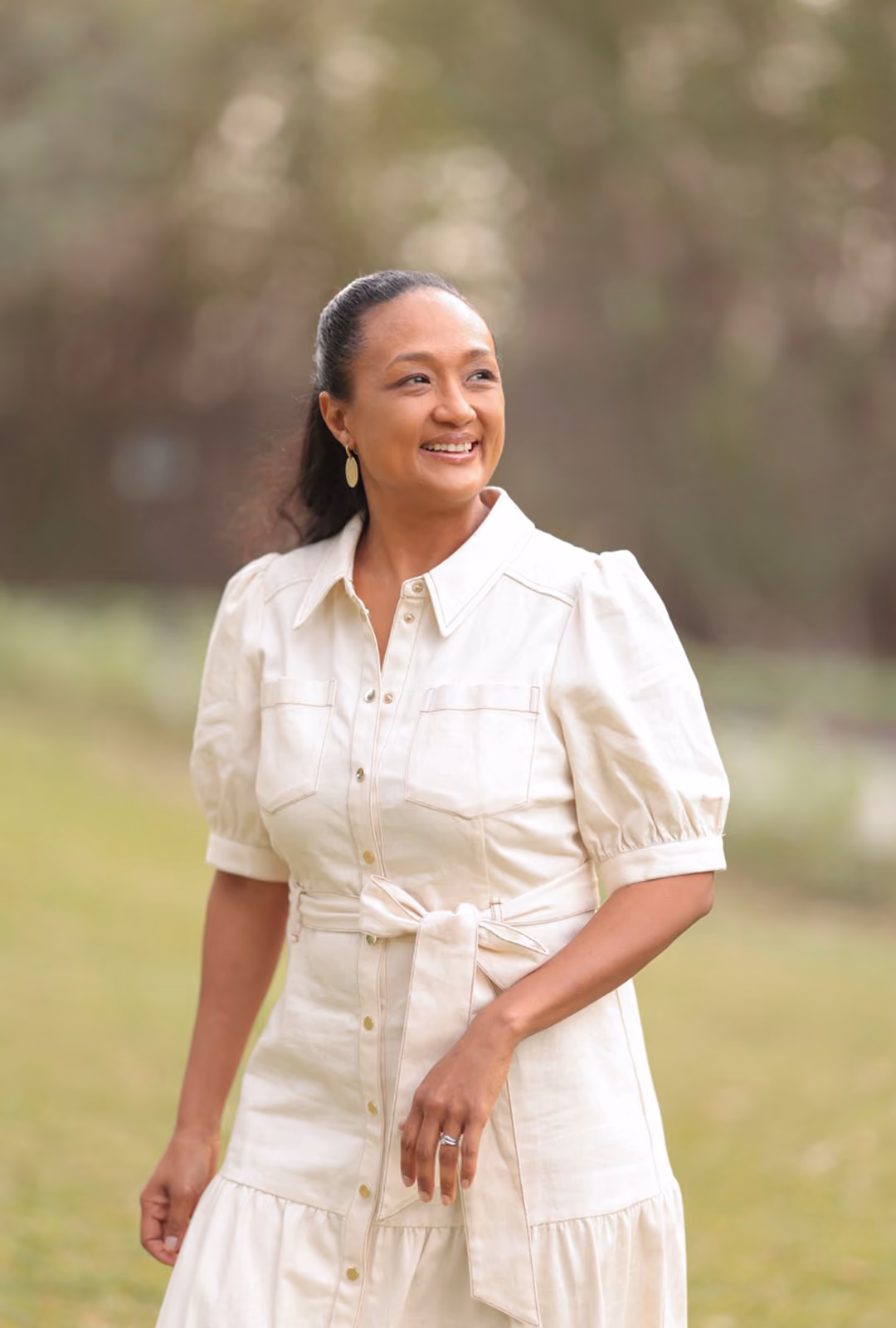 Clementine Clark wearing a light-colored button-up dress standing outdoors with blurred greenery in the background.