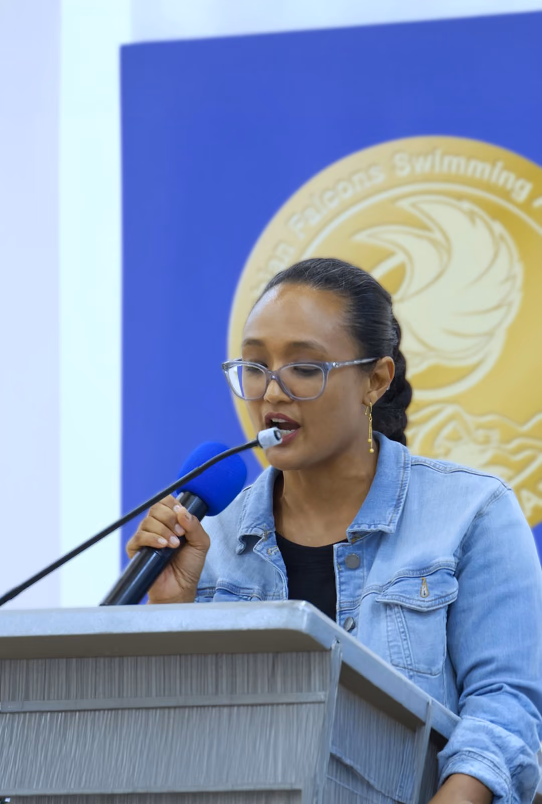 Clementine Clark speaking into a microphone at a podium, with a blue and gold emblem in the background.