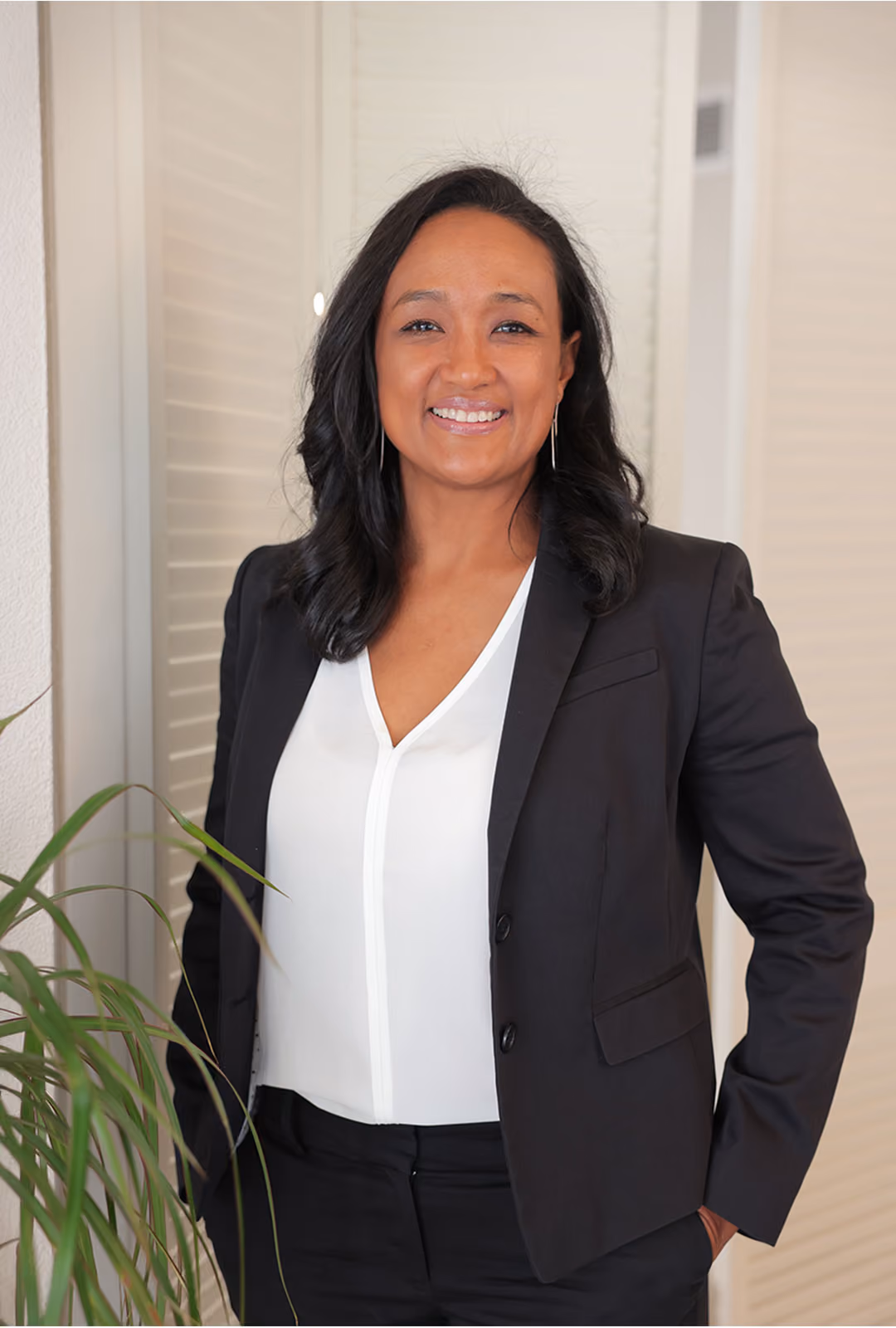 Clementine Clark wearing a black blazer over a white blouse, standing indoors next to a green plant with a light-colored background.