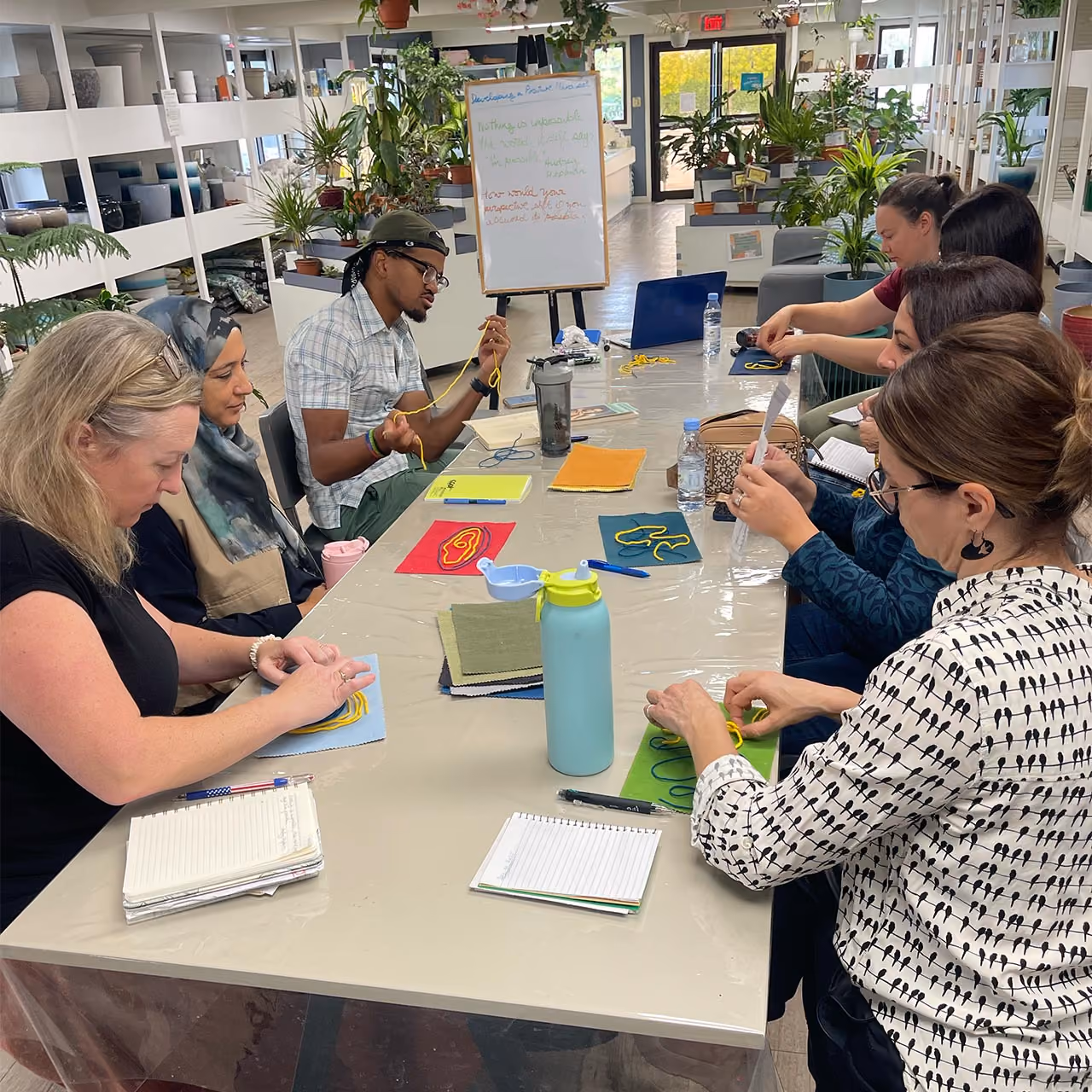 A group of seven diverse adults sitting around a rectangular table, engaging in a hands-on activity involving colored yarn and fabric swatches while taking notes and reading instructions.