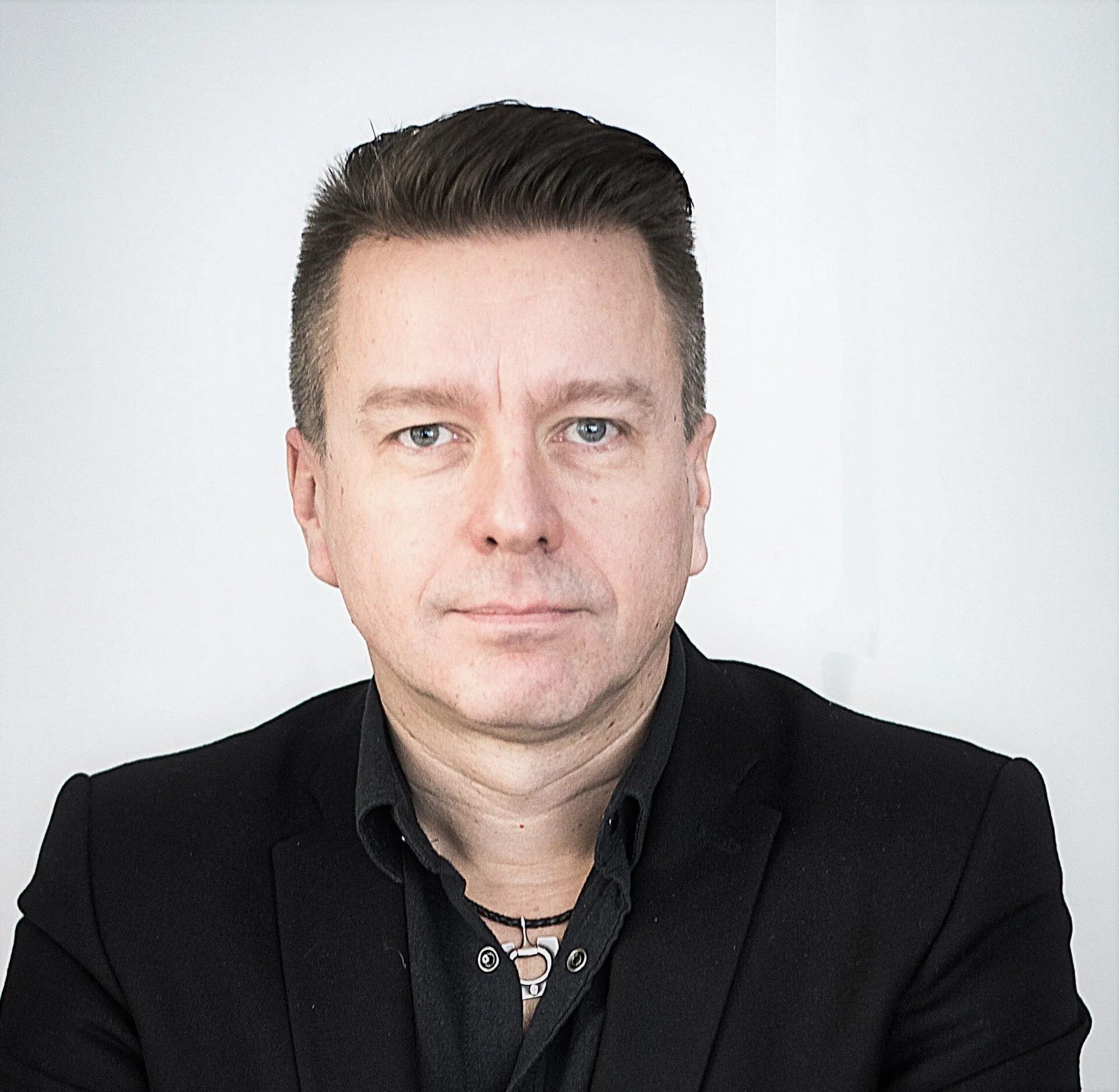Keski-ikäinen mies jolla on musta puku ja kaulakoru vaaleaa taustaa vasten.