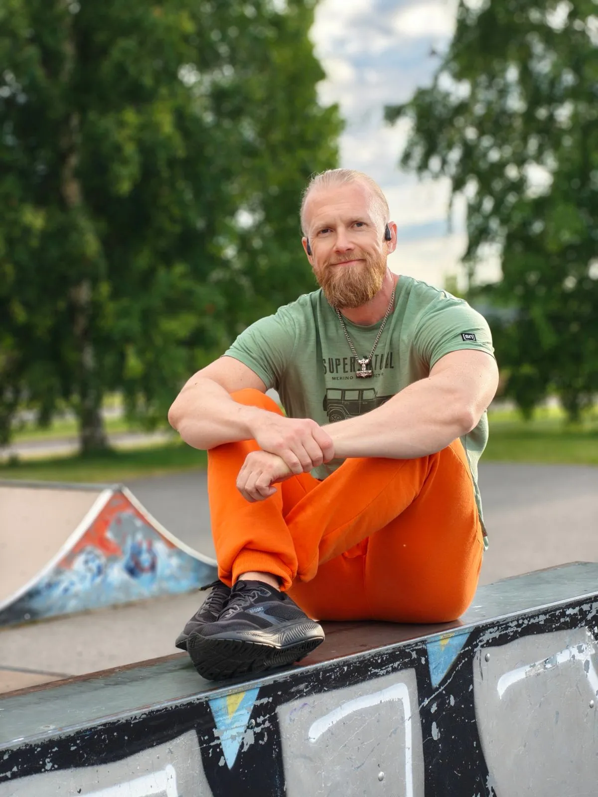 Vaalea, parrakas mies istuu ulkona skeittipuistossa vihreässä t-paidassa ja oransseissa housuissa.