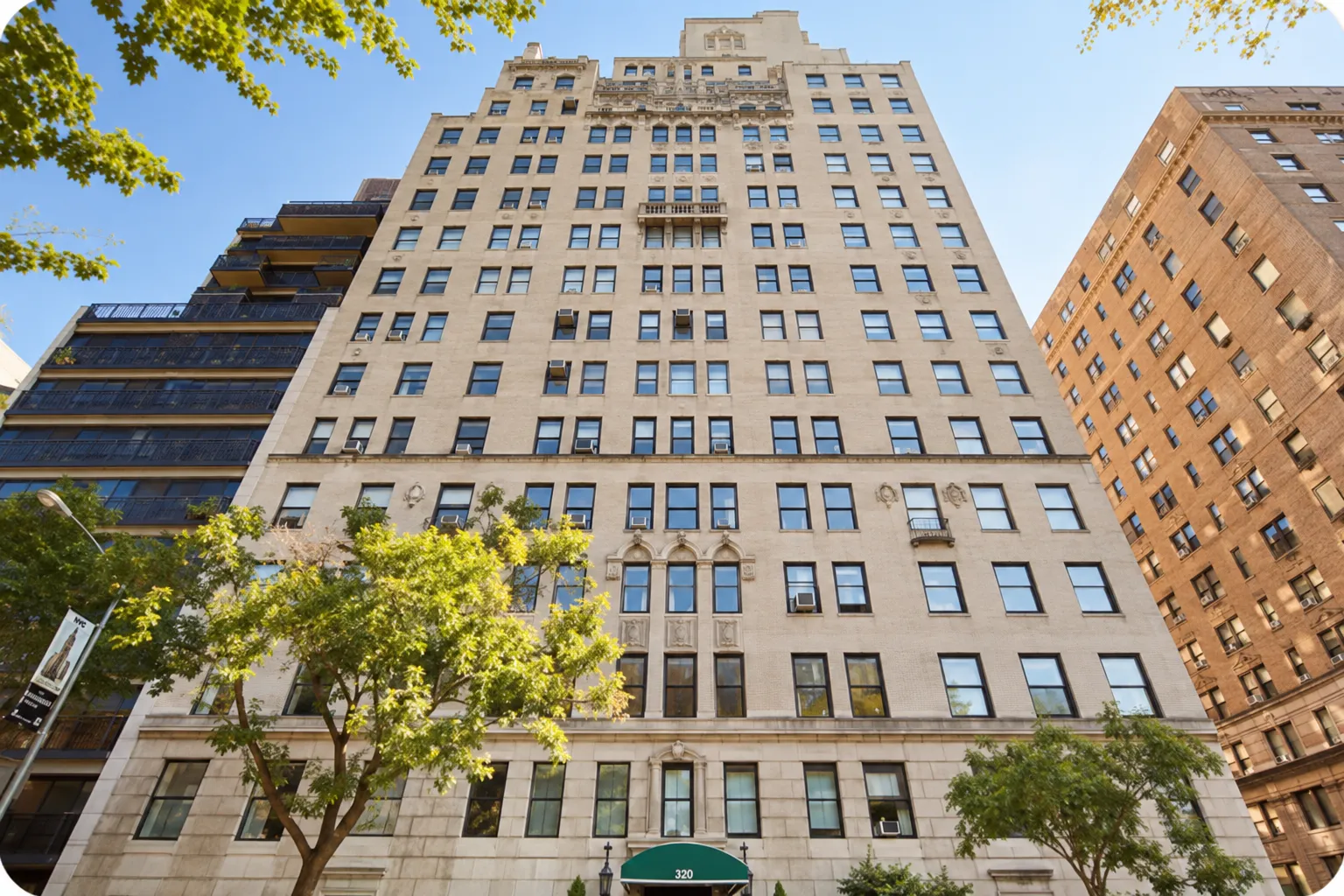 Tall beige apartment building with many windows, small balconies, and air conditioning units, flanked by two other buildings under a clear blue sky, with green trees in front and a green awning over the entrance displaying the number 320.
