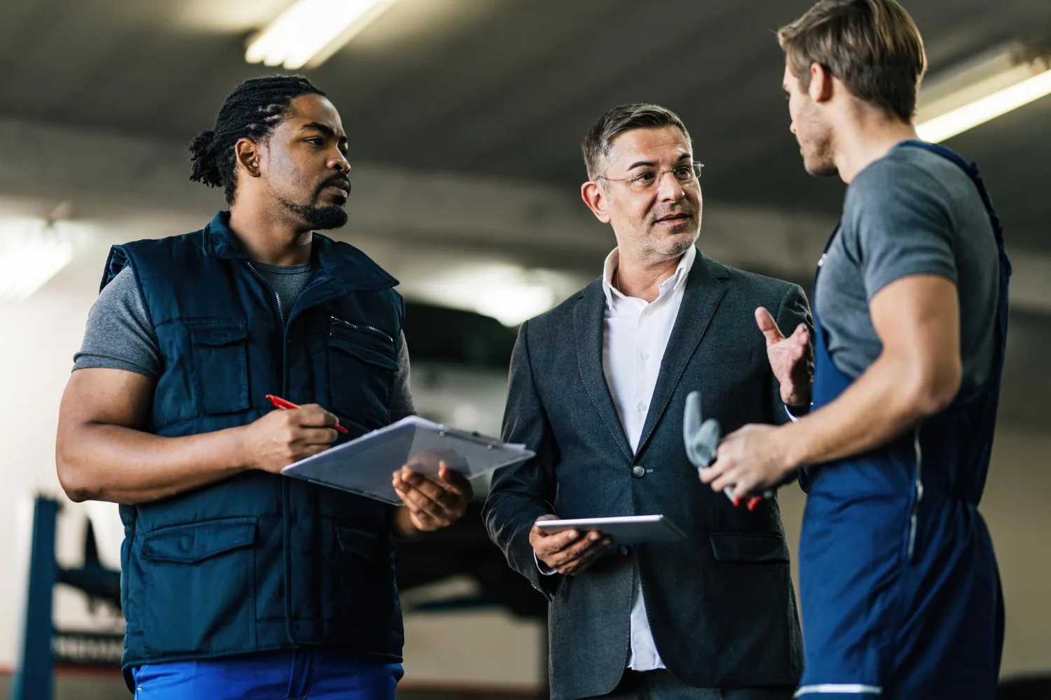 Three men in a workshop or garage discussing work; one in a suit holding a tablet, one in a blue vest holding a clipboard and pen, and one wearing a gray shirt and overalls holding gloves.