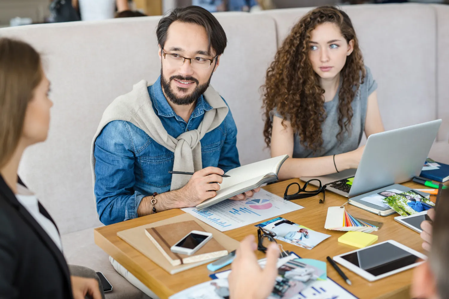 Three people in a meeting around a table with laptops, charts, photos, and notebooks; a man in a denim shirt and glasses holds a notebook and pen while speaking, a woman with curly hair listens attentively.