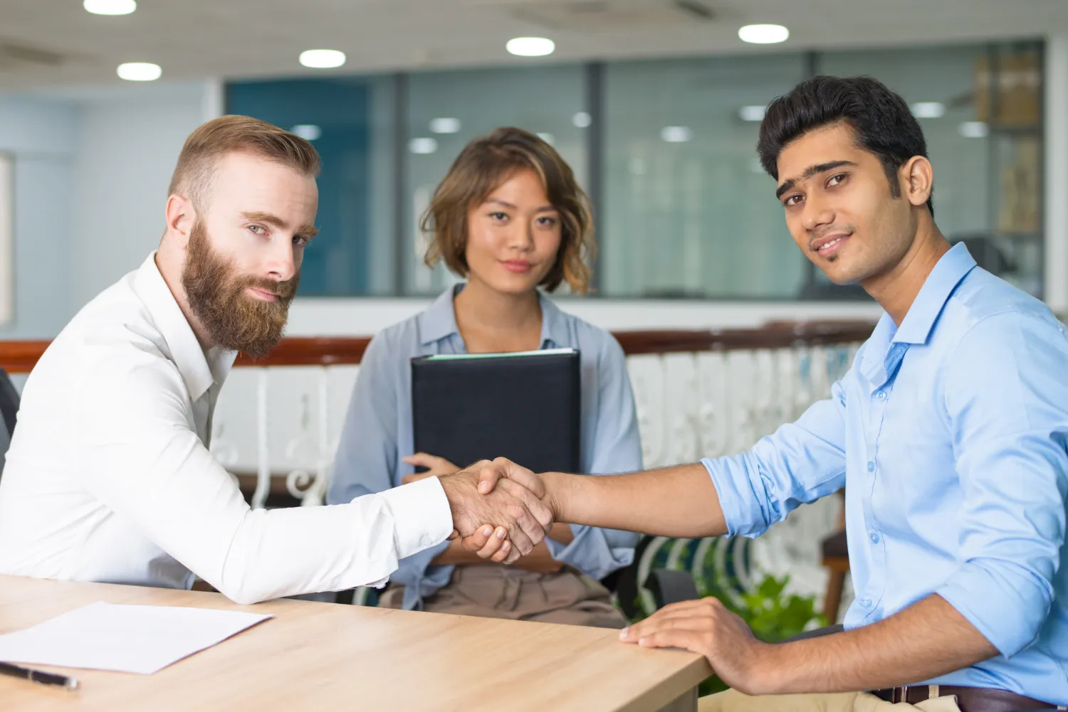 Two men shaking hands across a table in a professional setting while a woman holding a folder looks on, all three smiling slightly in a modern office environment.