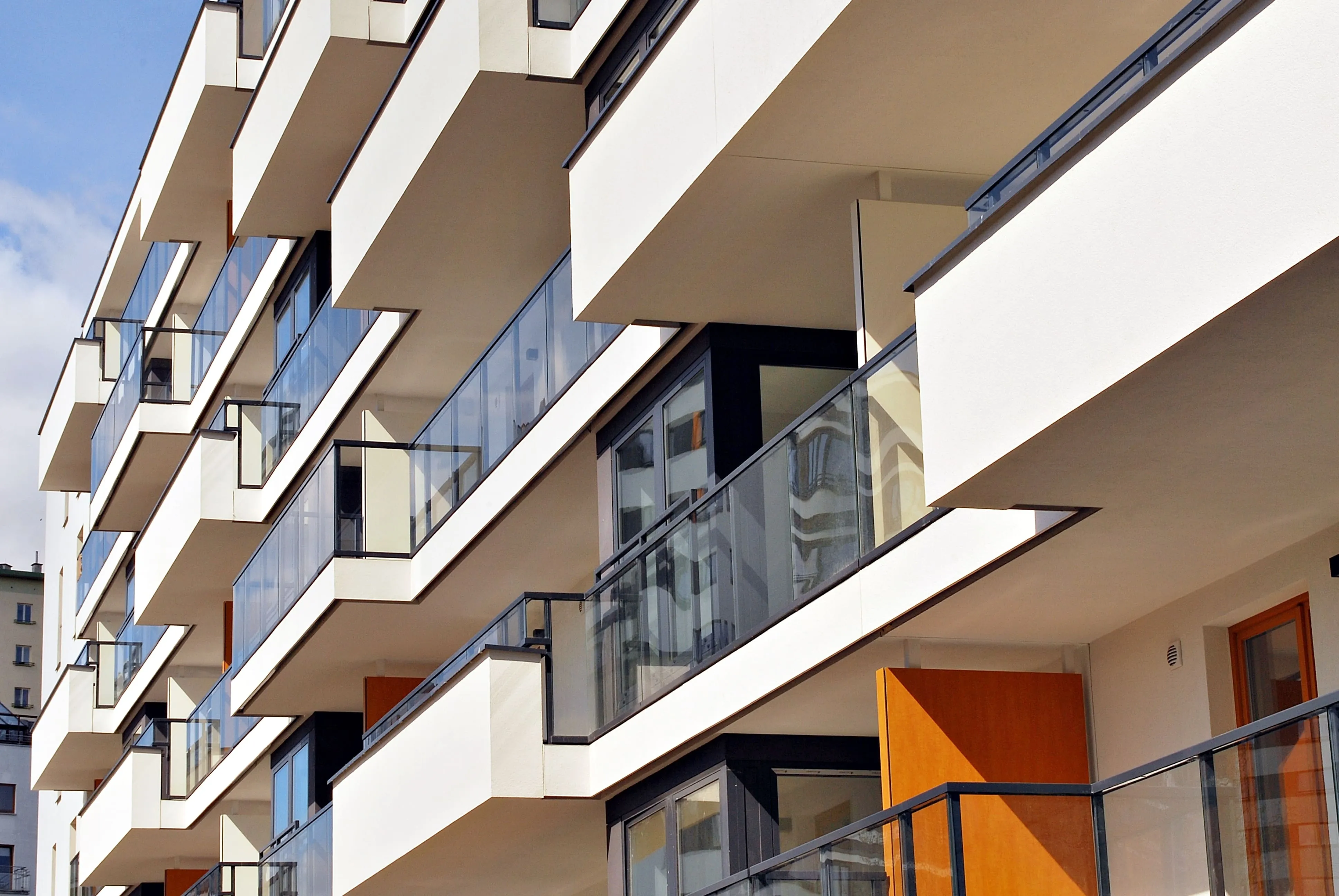 Modern apartment building facade featuring multiple balconies with glass railings and wooden doors, under a partly cloudy sky.
