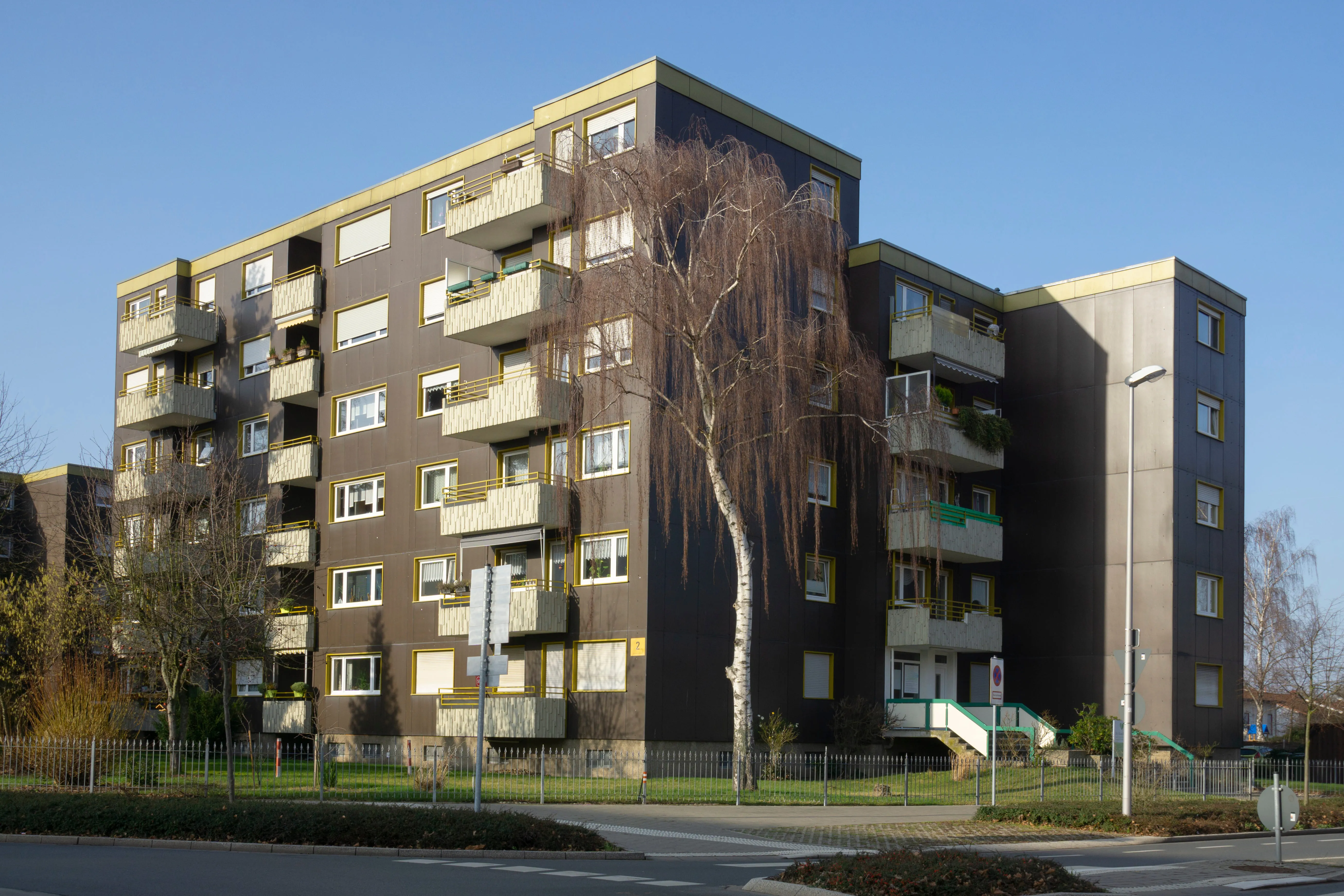 Four-story brown residential apartment building with pale yellow balconies and window frames, bare trees in front, fenced grassy area, and clear blue sky.
