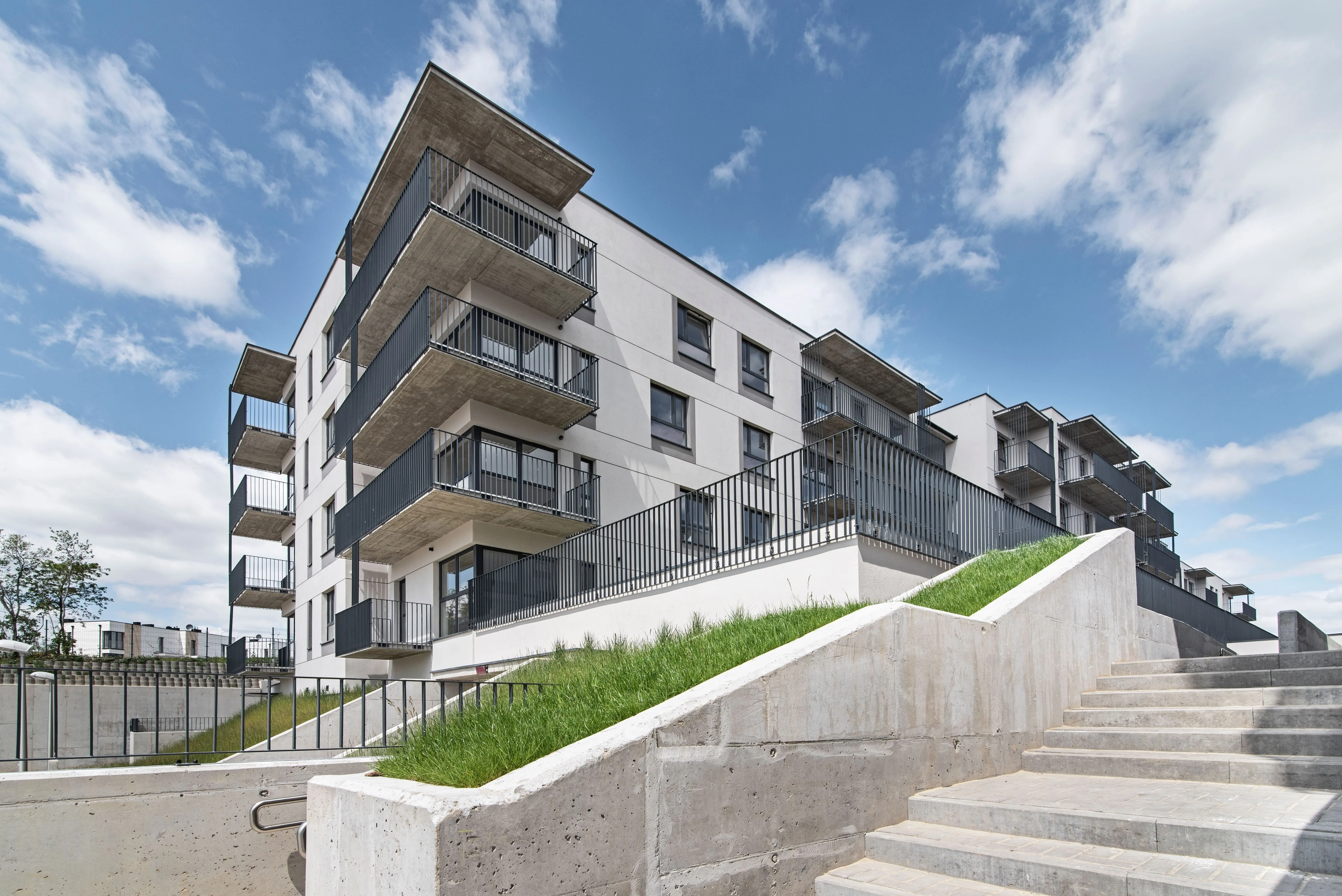 Modern multi-story apartment building with multiple balconies and black metal railings, white walls, concrete accents, and stairs leading up to the entrance under a partly cloudy blue sky.