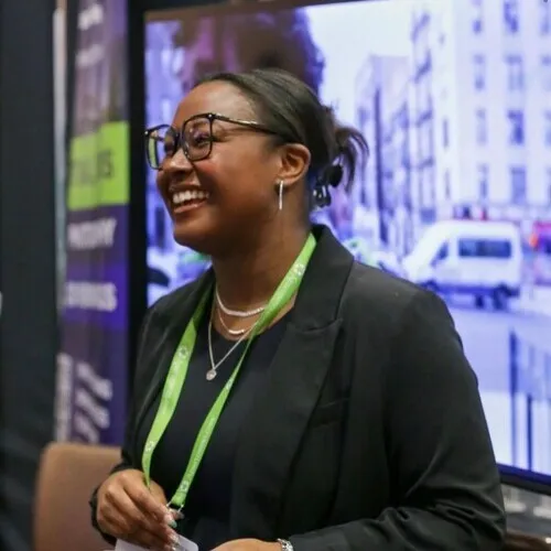 Smiling woman wearing glasses and a black blazer with a green conference lanyard and name badge, standing in front of a blurred screen displaying a city scene.
