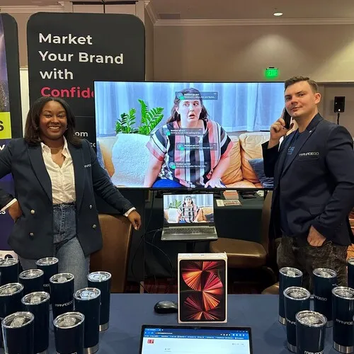 Two people standing at a booth with promotional tumblers, a tablet, and a laptop on the table, with a large screen behind them showing a woman sitting on a couch using sign language, and text overlay for accessibility.