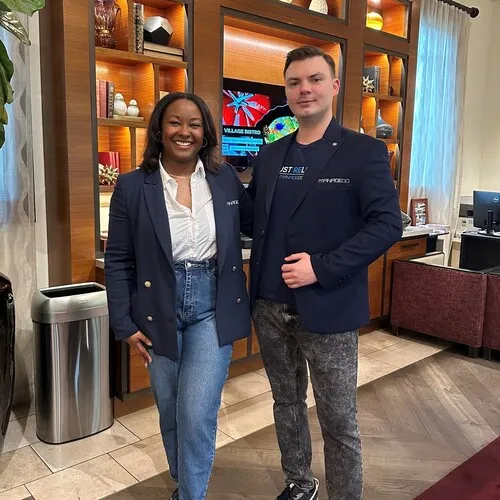 A man and a woman standing side by side indoors in front of a wooden bookshelf with decorative items and a TV screen, both wearing dark blazers; the woman has on jeans and a white shirt, while the man wears patterned pants and a black t-shirt.