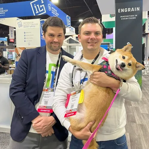 Two men at a trade show booth; one man with a badge labeled 'exhibitor' is holding a happy Shiba Inu dog wearing a pink harness and leash, while the other man stands beside him. The booth displays signs for Interior Logic Group and Engrain.