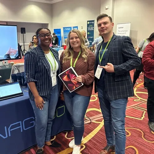 Three people standing and posing at an indoor event with conference name badges and green lanyards; two women and one man, the woman in the middle holding a tablet, with display tables and other attendees in the background.