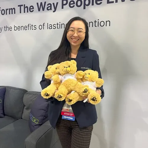 Smiling woman wearing glasses and a dark blazer holding three plush yellow teddy bears with paw prints on their feet, standing indoors near a gray couch and white wall with partial text in the background.