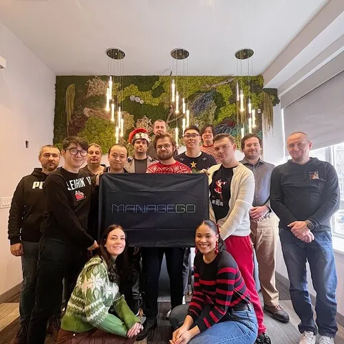 Group of fourteen people posing indoors, holding a black banner with 'MANAGEGO' logo, standing and kneeling in front of a green moss wall with hanging lights.
