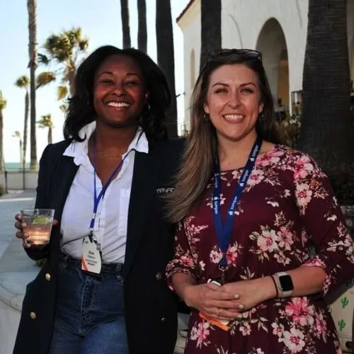 Two smiling women standing outdoors near palm trees and a building with arches; one woman holds a drink and wears a dark blazer over a white shirt with jeans and a lanyard, the other wears a floral dress with a lanyard and smartwatch.