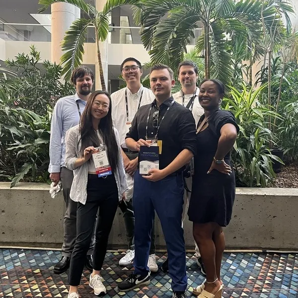 Group of five diverse adults standing indoors in front of lush tropical plants, smiling at the camera, with some wearing conference badges.