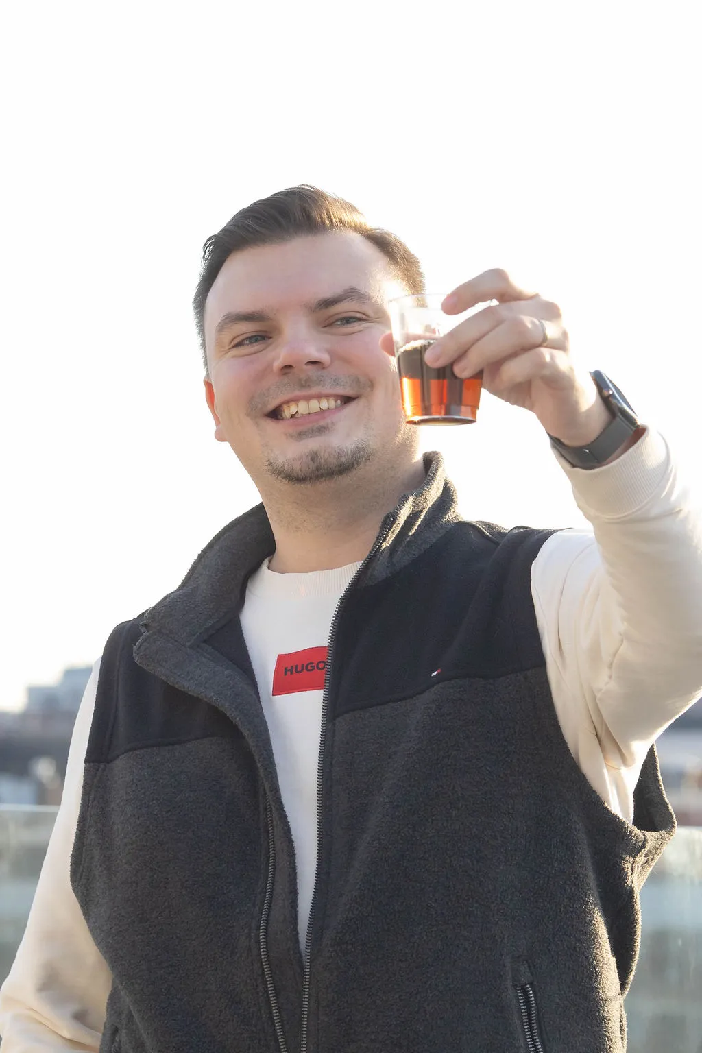 Smiling man wearing a dark fleece vest and a light-colored long sleeve shirt raising a small glass with a dark beverage in a toast.