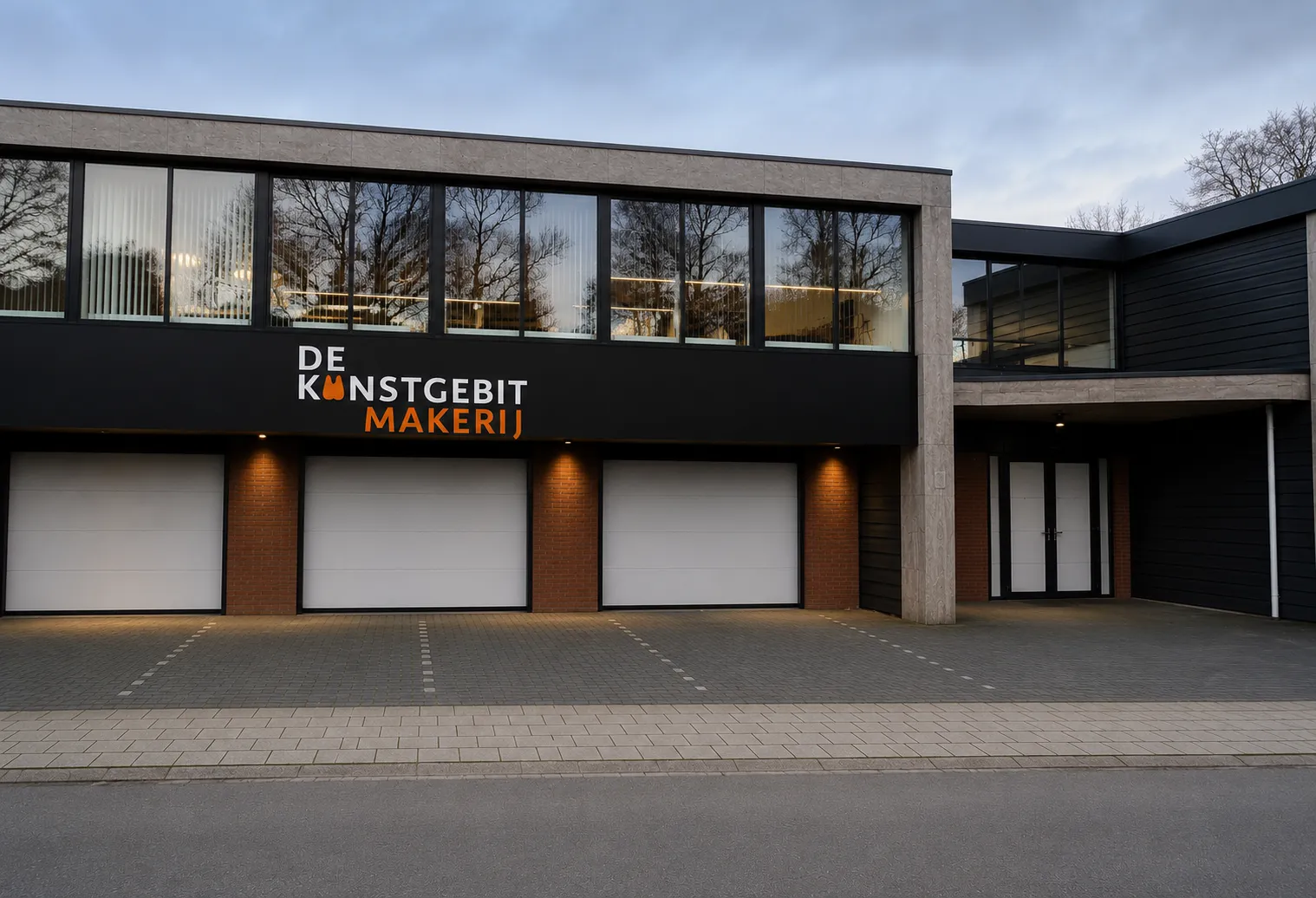 Modern building with three white garage doors and large windows above reflecting trees, with signs reading DE KUNSTGEBIT MAKERIJ in white and orange letters on a black panel.