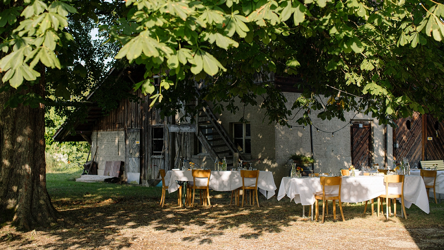 Aussentische mit weißen Tischdecken und Holzküchenstühlen unter Schatten grosser grüner Bäume vor einem alten rustikalen Gebäude mit Holz- und Putzfassade, vorbereitet für ein Sommerevent.
