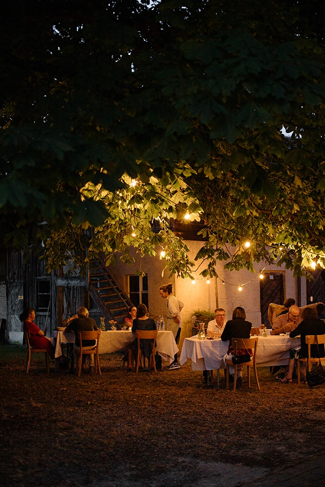 Menschen sitzen abends unter einem grossen Baum an  gedeckten Tischen mit weissen Tischdecken, beleuchtet von warmen Lichterketten, vor einem Haus mit Holzstiegen und einer Holztür.