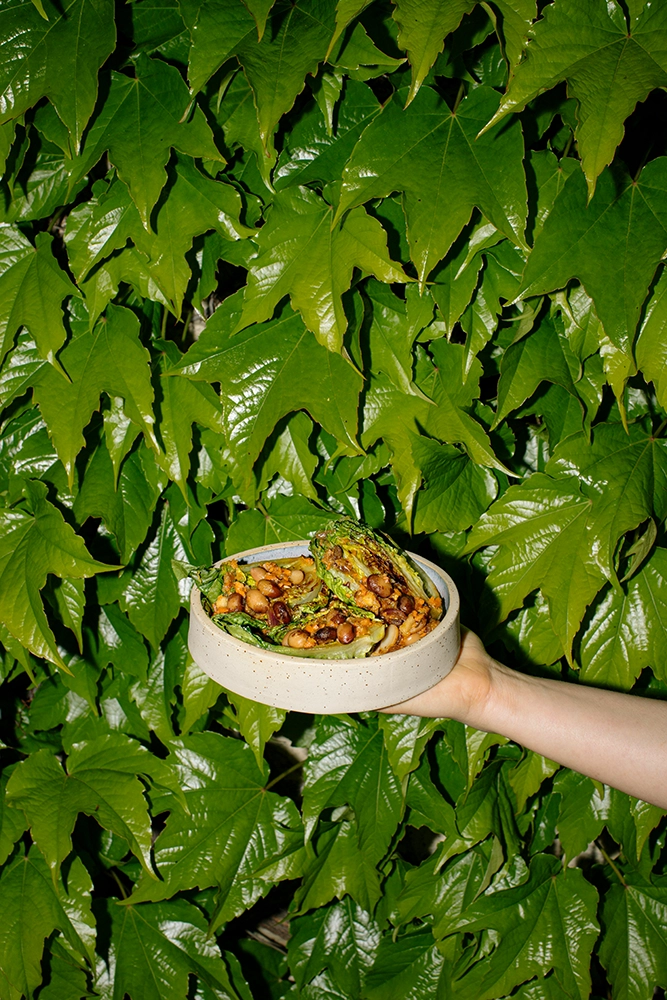 Michaela Frank hält eine Keramikschüssel mit einem Salat aus gegrilltem grünen Gemüse und Kichererbsen vor einem Hintergrund aus grünen Blättern.