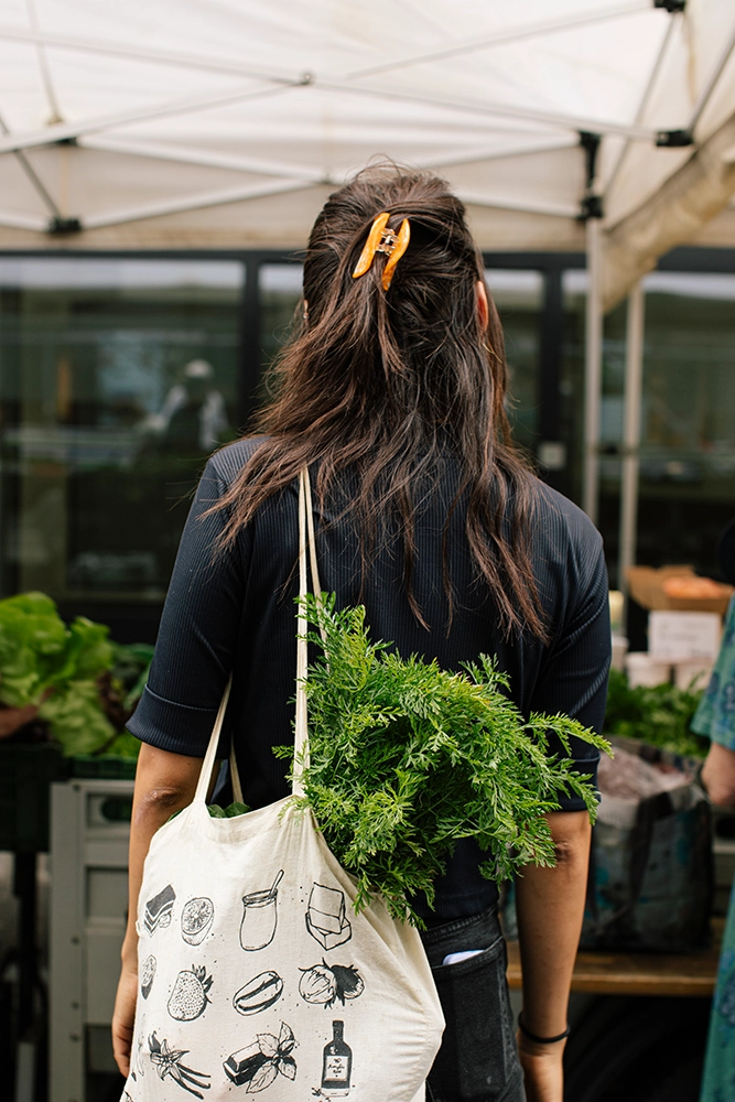 Michaela Frank trägt eine bedruckte Stofftasche mit frischen Karottengrün besucht einen Markt.