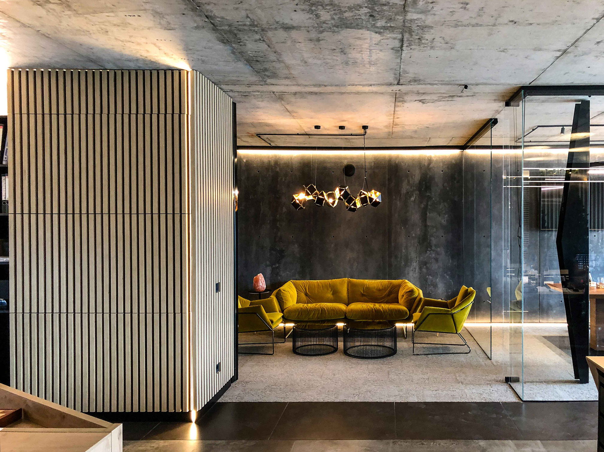 The studio — mustard seating and a faceted pendant under exposed concrete, beside a slatted timber volume