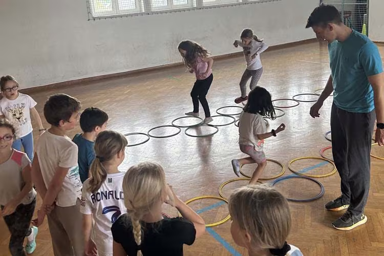 Otroci skačejo po obročih na tleh telovadnice, trener jih spremlja