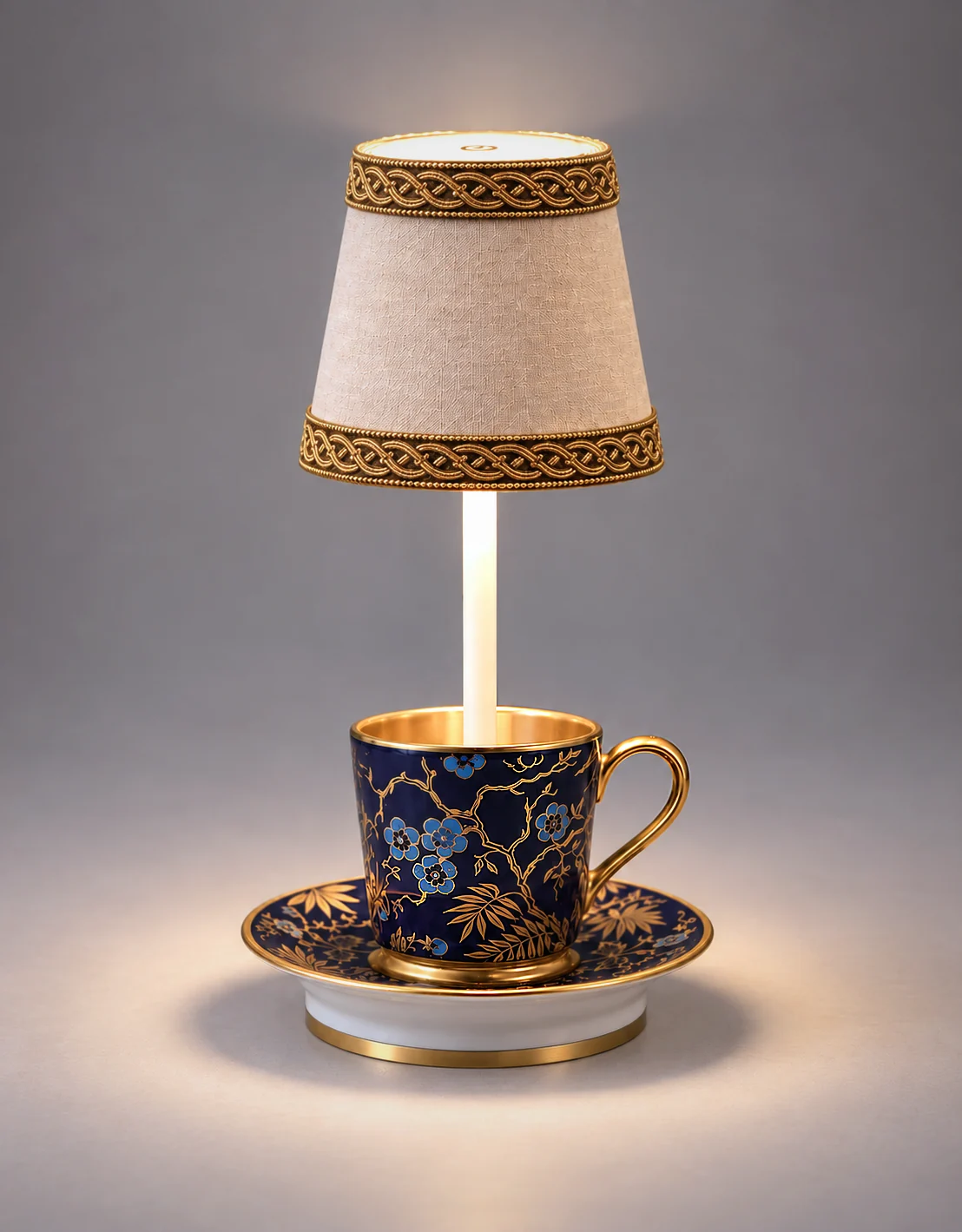 Table lamp with a small beige lampshade decorated with lace on top of a dark blue teacup with gold and blue floral patterns, sitting on a matching saucer on a marble surface.