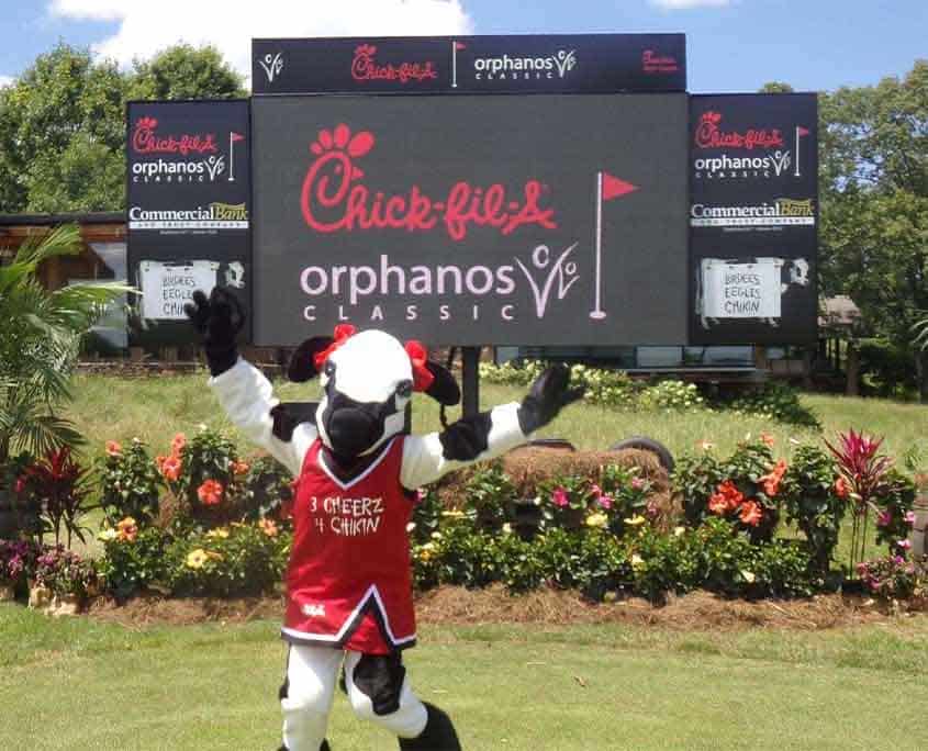A cow mascot in a red and white jersey stands in front of a large Chick-fil-a sign.