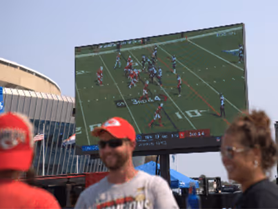 Outdoor large screen showing a football game with people watching in the foreground.