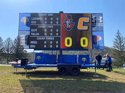 Outdoor scoreboard on trailer showing zero scores for courts one, two, and three during a daytime event.