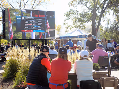 People sitting outdoors watching a large screen showing sports event details during a sunny day.