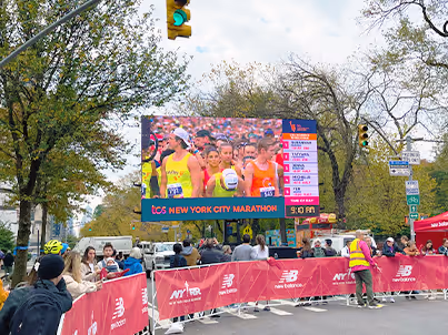 Outdoor crowd watching live coverage of runners in New York City Marathon on large screen.