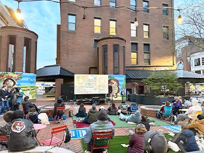 People sitting on chairs and blankets outdoors watching a screen in a courtyard with murals on brick walls.