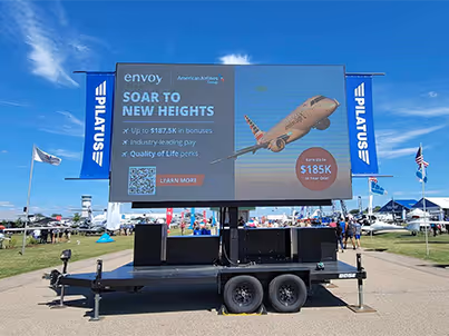 Digital advertising display on trailer at outdoor air show with planes and clear sky in background.