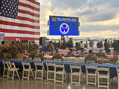 Military personnel seated and standing in hangar with large American flag and William Tell 2023 event screen.
