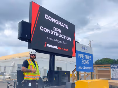 Man in safety vest and helmet stands beside digital sign congratulating DPR Construction at a PPE free zone.