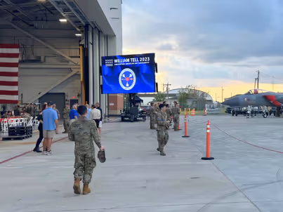 Military personnel walk near aircraft and a screen displaying 'William Tell 2023' inside an airbase hangar.