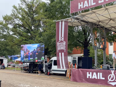 Outdoor stage with 'Tailgate' banner and large screen showing a music performance in a park setting.