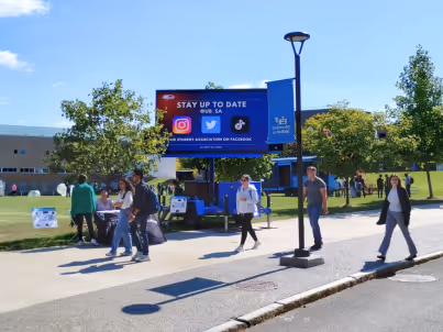 People walk near a digital signboard on campus promoting staying updated on social media.