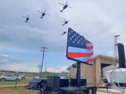 Six helicopters flying in formation above a trailer-mounted large flag resembling the US flag.
