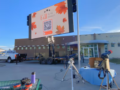 Outdoor scene with a large digital sign displaying fall leaves and a QR code near telescopes on tripods.